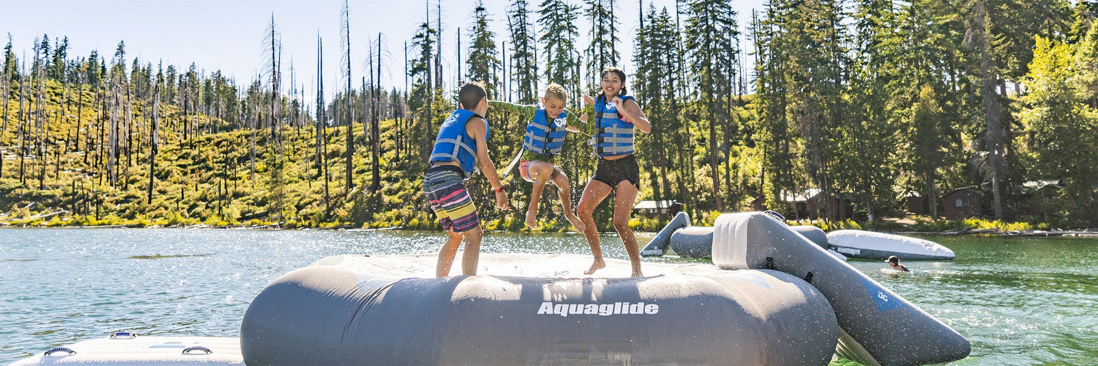 Three kids in life jackets jump on an Aquaglide water trampoline on a lake surrounded by trees.