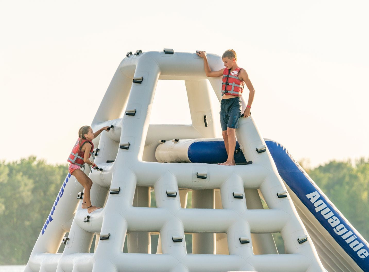 Two kids wearing life vests climb a large inflatable water obstacle on a lake.