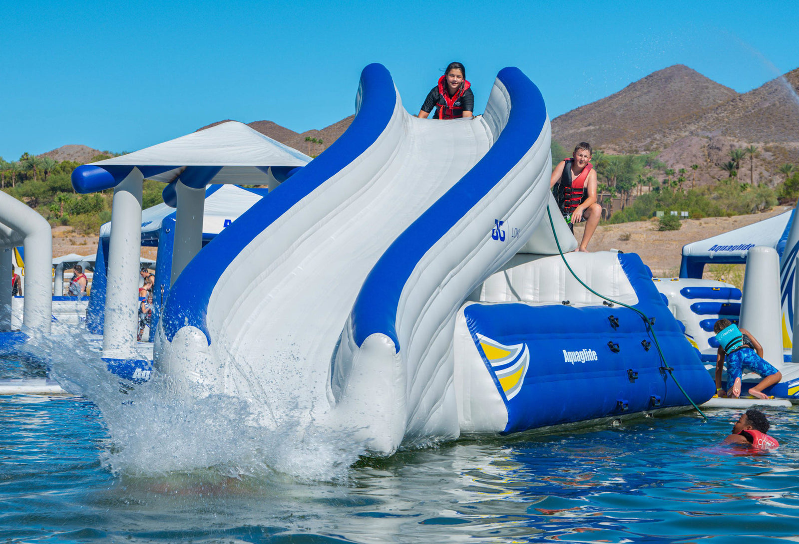 People sliding down an inflatable water slide into a lake with mountains in the background.