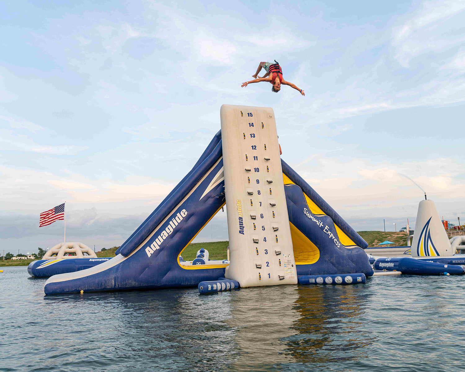 Person flipping off an inflatable climbing wall in a water park under a cloudy sky.