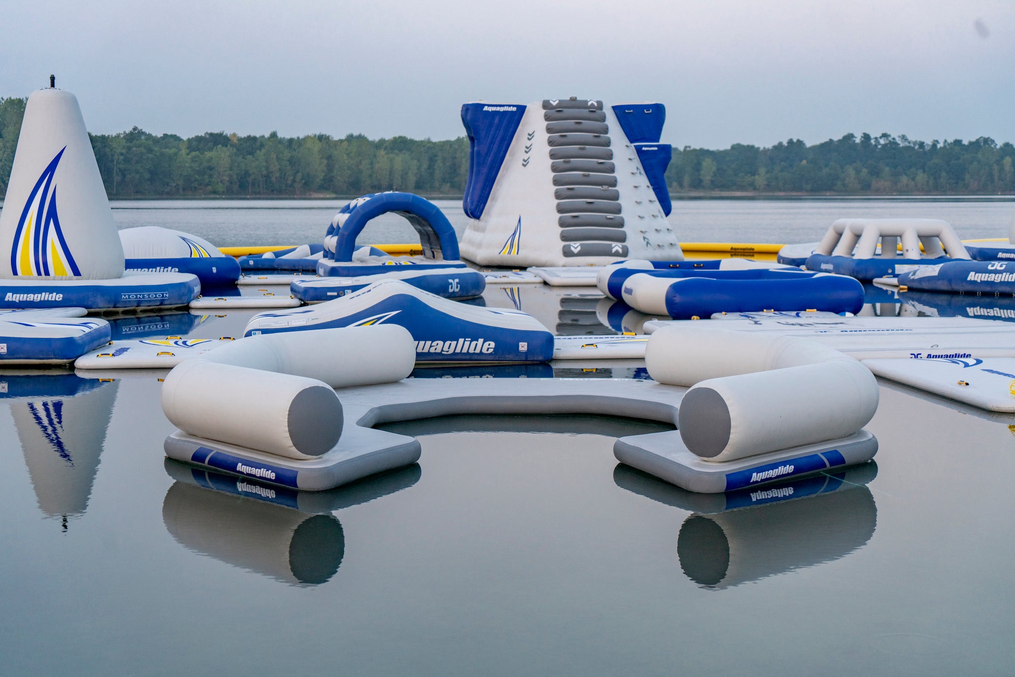 Aquaglide lounge float faces a tall climbing tower in a calm open-water park setting.