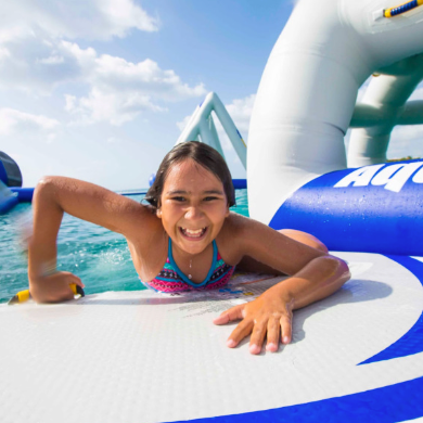 Girl in a swimsuit smiling while climbing inflatable structure in blue water.