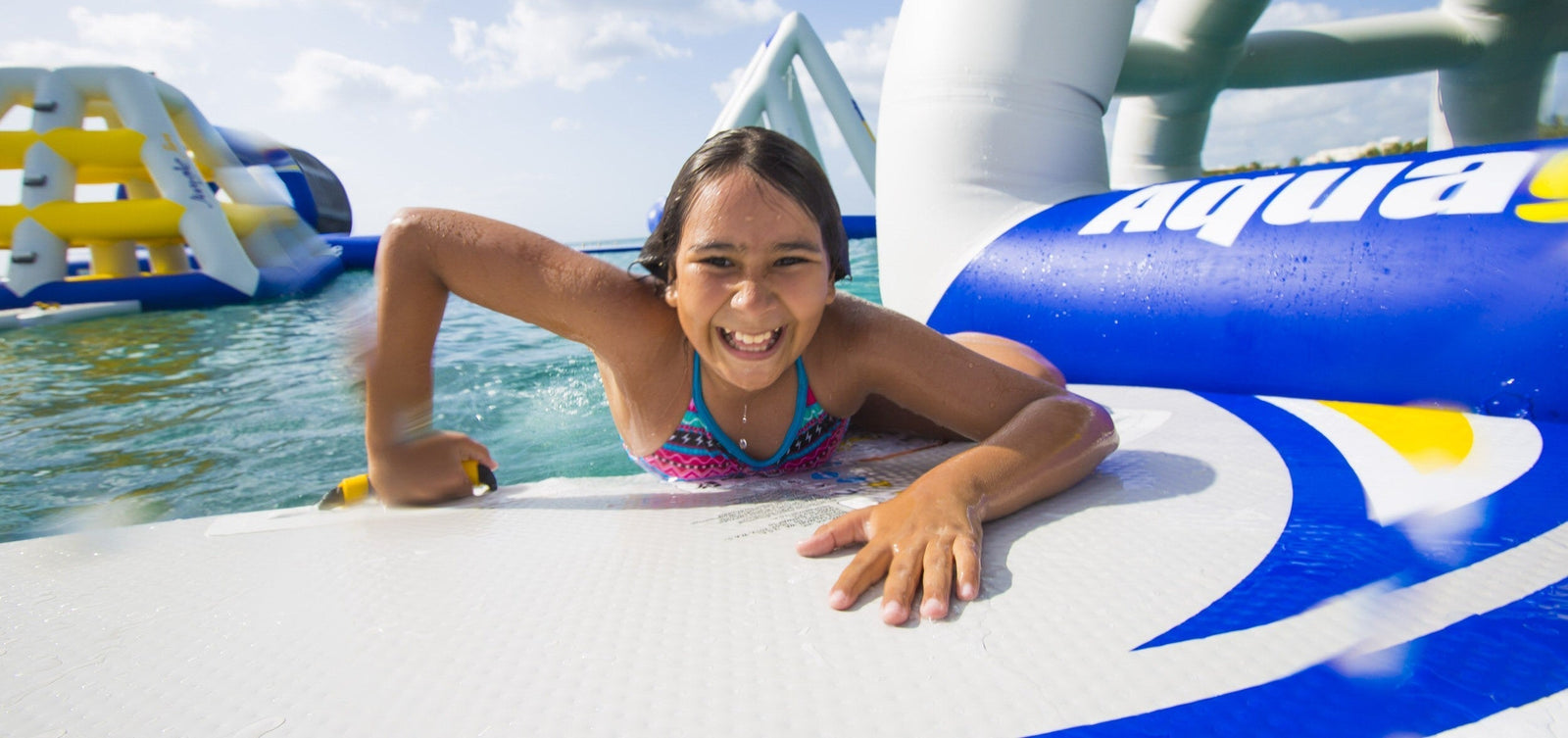 Child smiling while climbing onto a floating inflatable obstacle at a water park.