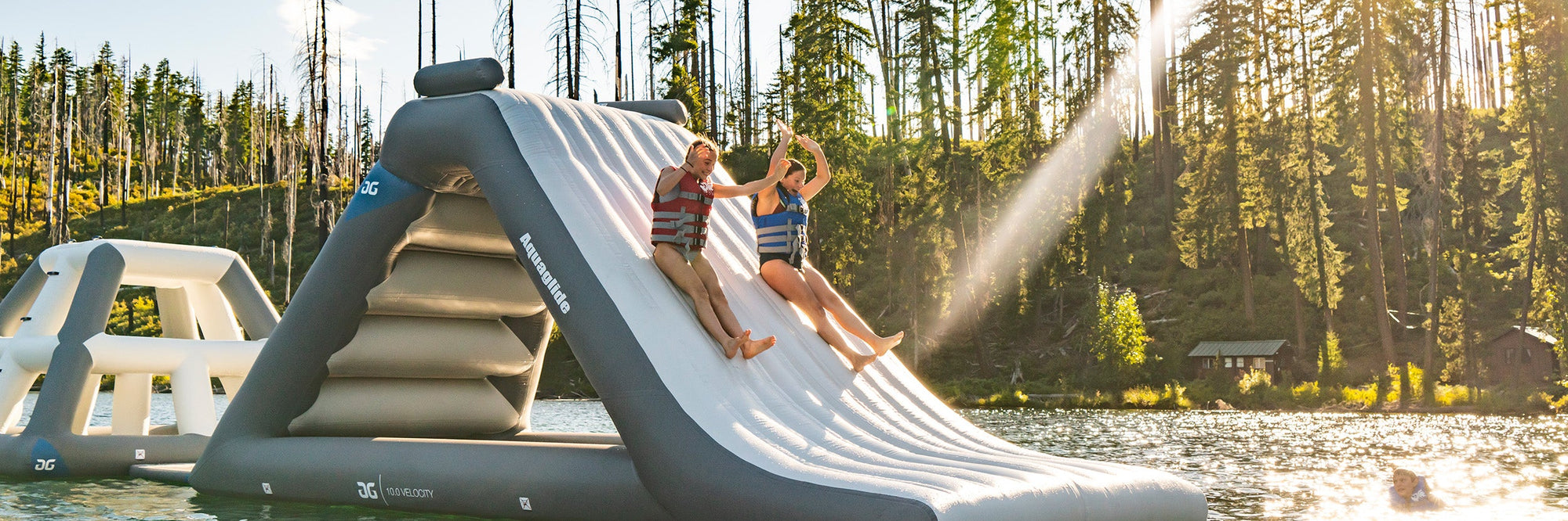 Two people sliding down an inflatable water slide into a lake, surrounded by trees and cabins under a sunny sky.