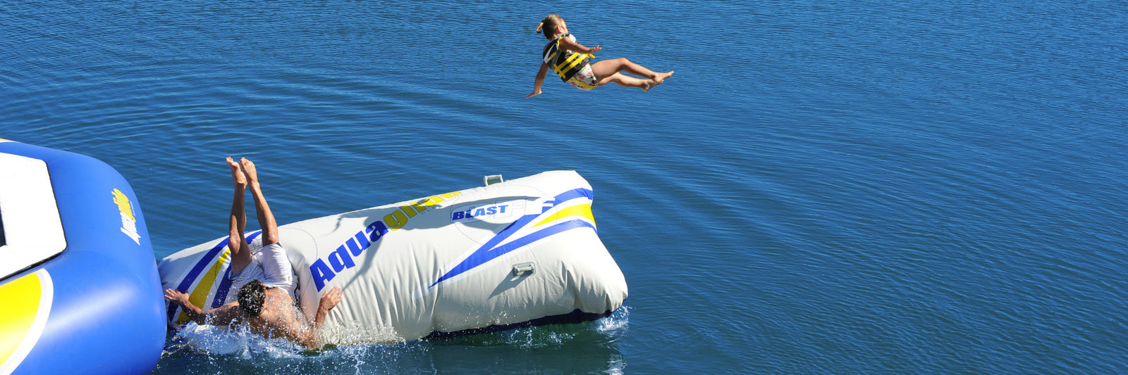 Child launched into air from inflatable blob on lake, another person in water; blue and white water toys visible.