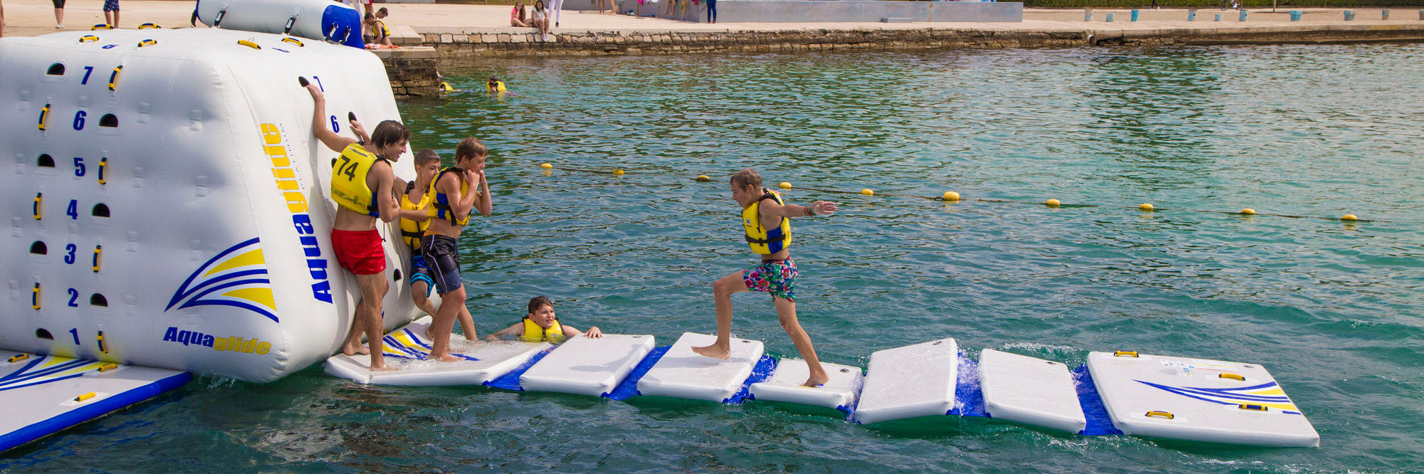 Children on a floating obstacle course in a lake, wearing life jackets and stepping on foam pads.
