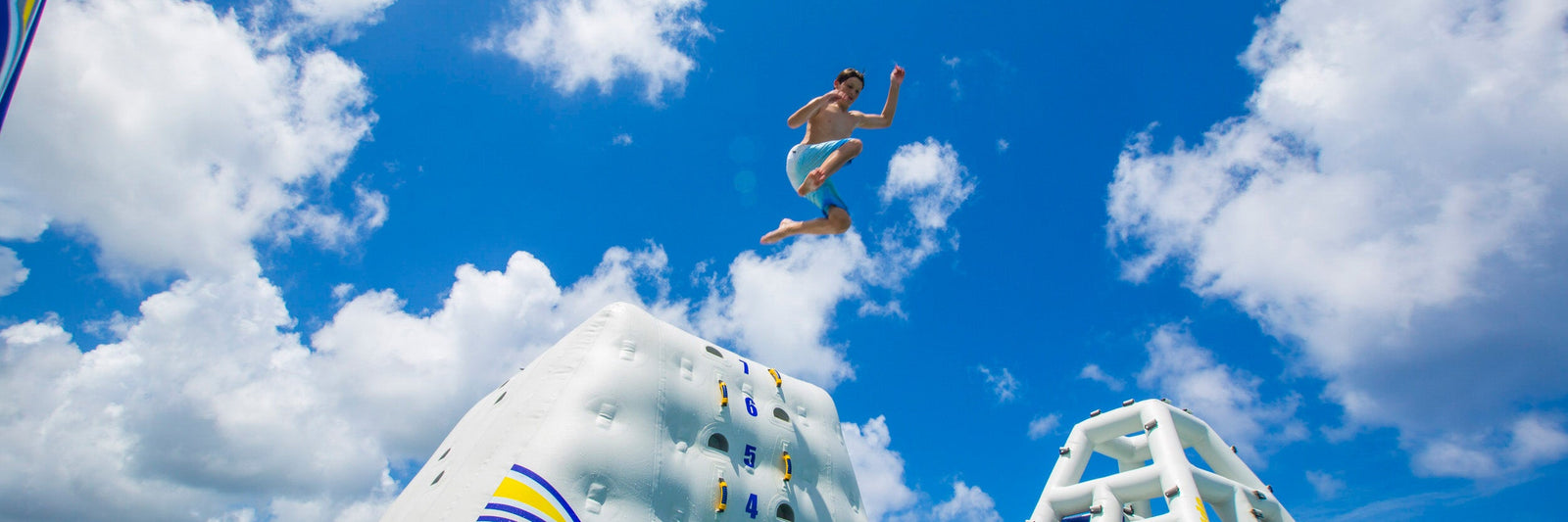 Person in blue shorts jumps from inflatable climbing wall against a bright blue sky with fluffy clouds.