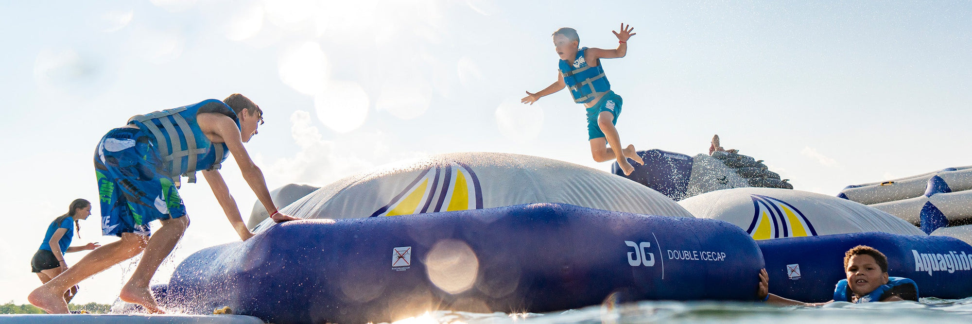 Children playing and jumping on inflatable water park equipment in the sun, wearing life jackets.