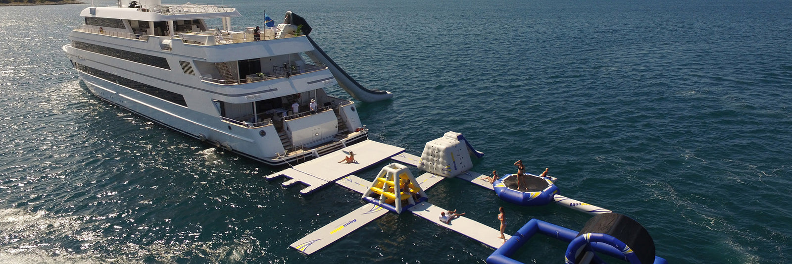 Large yacht with a water slide and floating inflatable play area on a sunny day at sea.