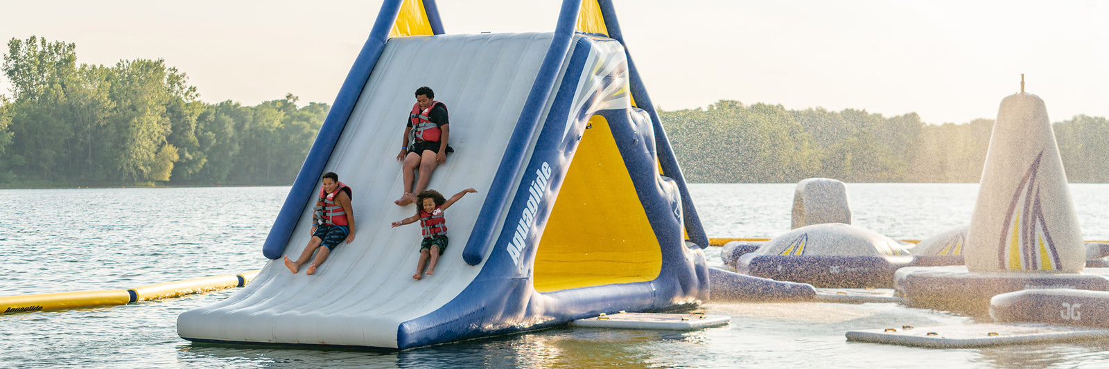 Three people in life vests slide down a large inflatable slide into a lake, with inflatable obstacles in the background.