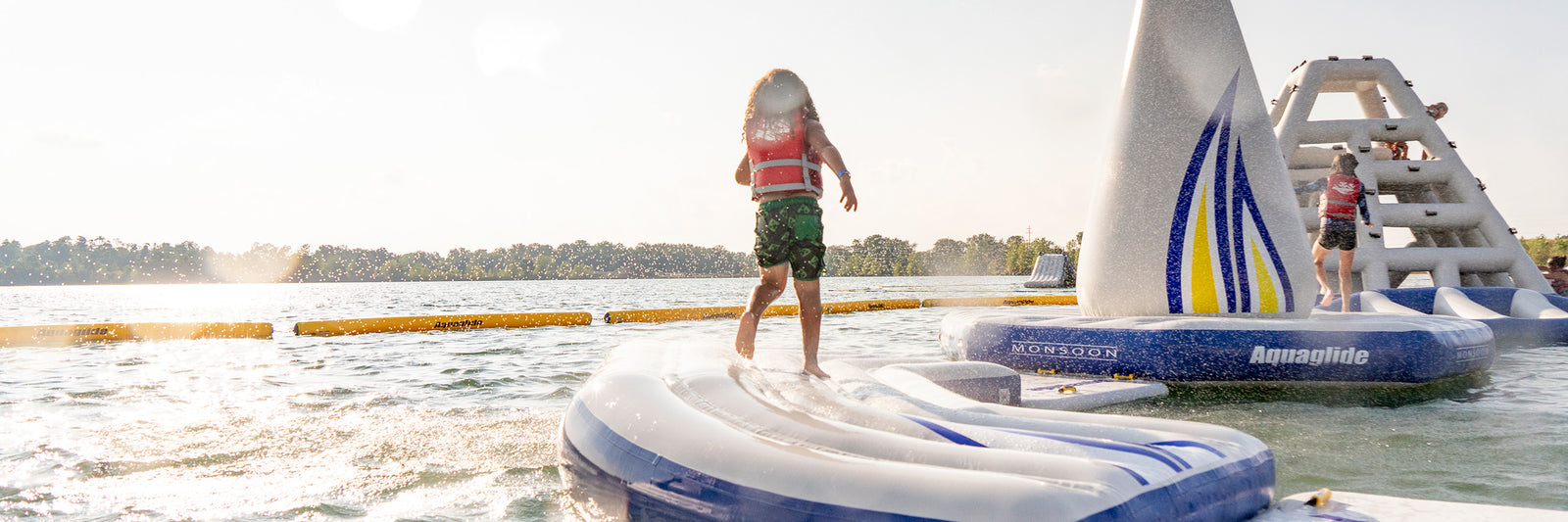 Child in a life jacket runs on a floating water obstacle course in a sunny lake.