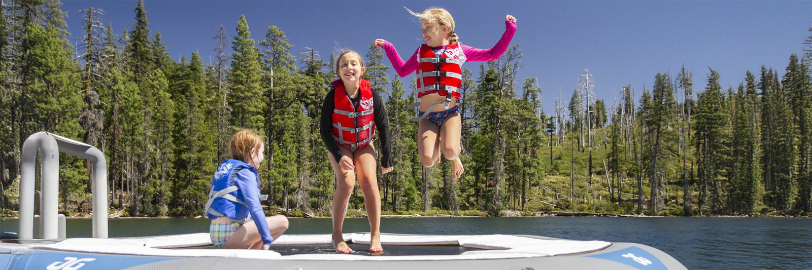 Three kids in life jackets joyfully jump on a large inflatable by a scenic lake with a forested background.