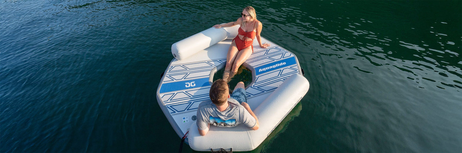 Two people relax on a floating inflatable platform on a calm lake.