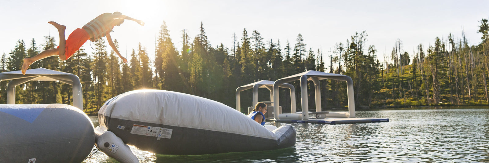 Person leaping onto inflatable raft on a lake, surrounded by trees and floating docks, under a clear sky.
