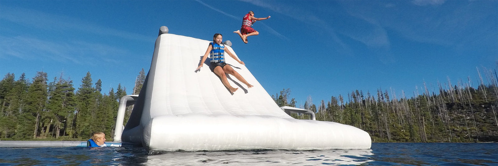 People in life jackets enjoying a large inflatable slide on a lake with a forested shoreline.