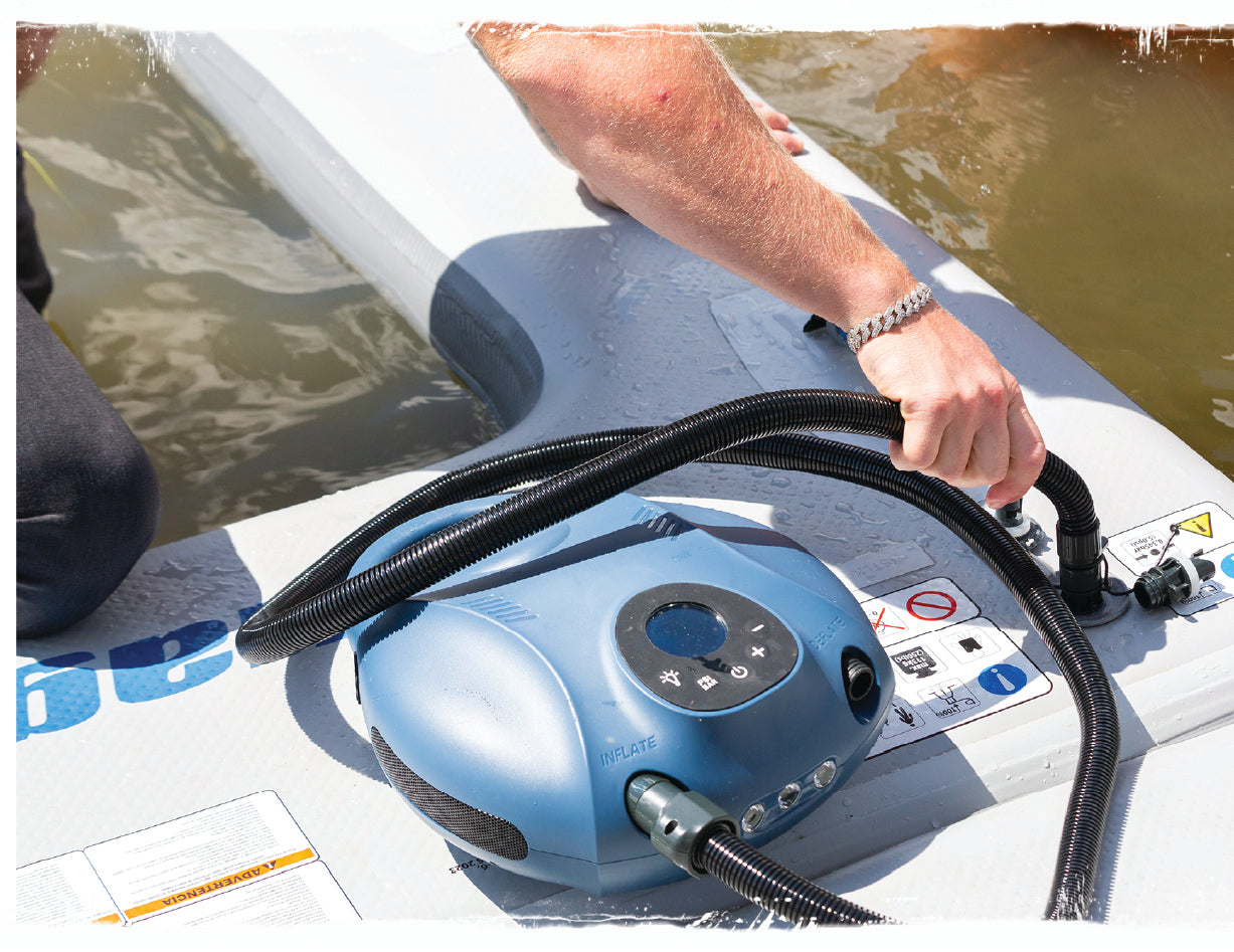 Close-up of person connecting electric pump hose to inflatable platform at water’s edge.