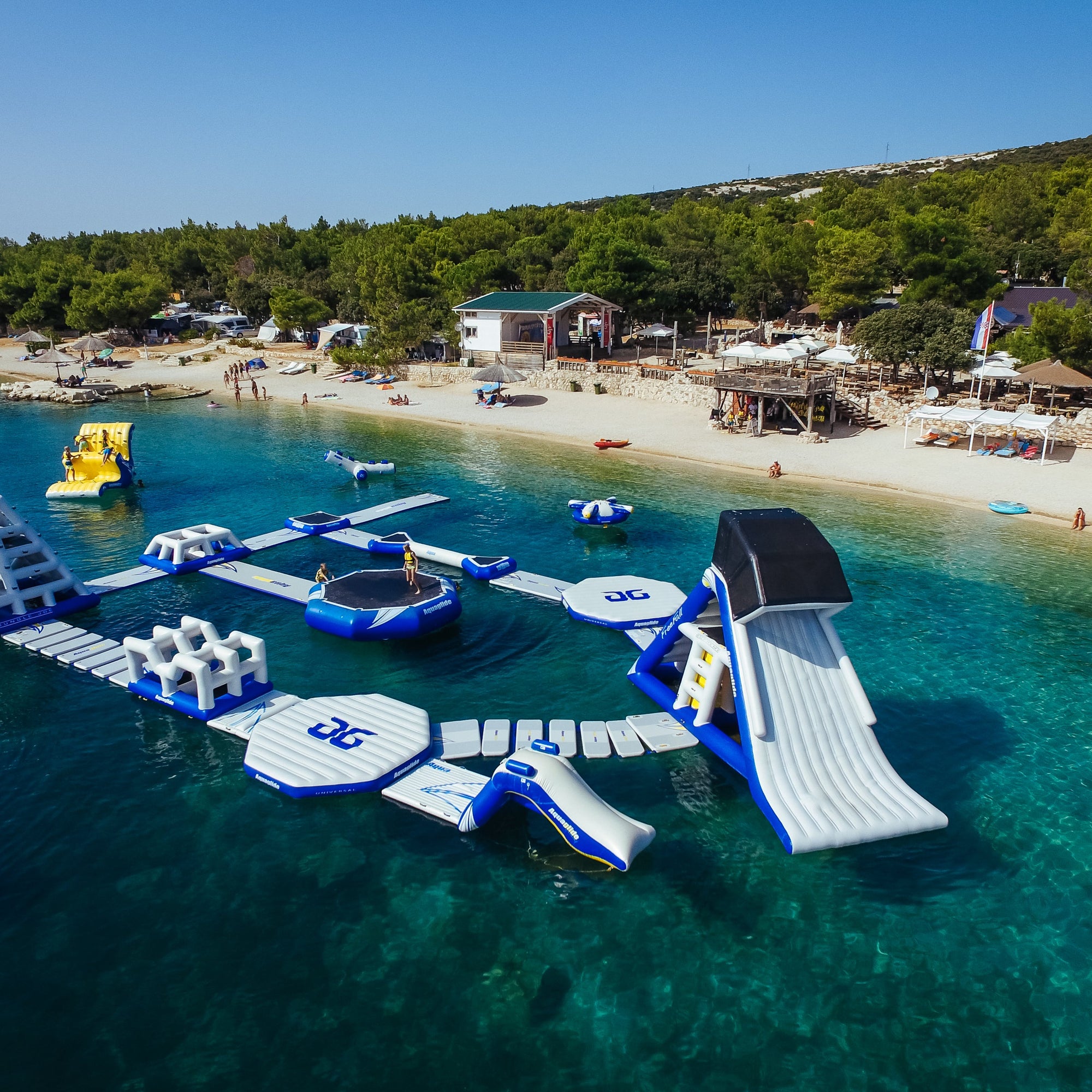 Large Aquaglide floating water park bear the shore of a lake with multiple kids playing on the slides and other obstacles.