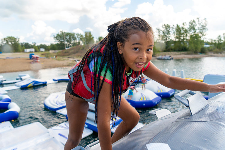 Child climbing an inflatable water structure, wearing a life vest, with trees in the background.