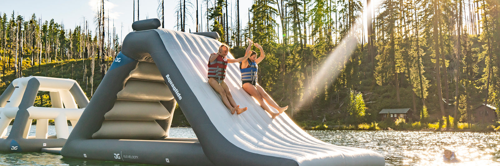 Two people slide down a large inflatable slide into a lake on a sunny day.