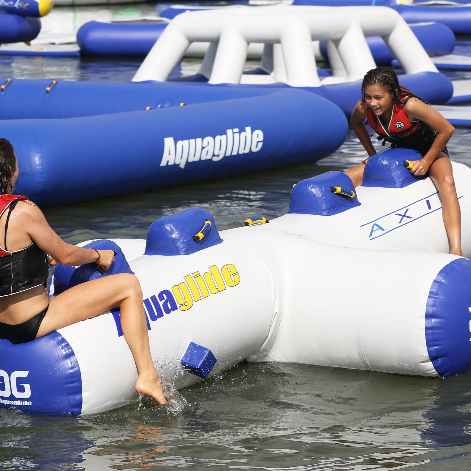Two people on an inflatable water toy, wearing life jackets, on a sunny day.