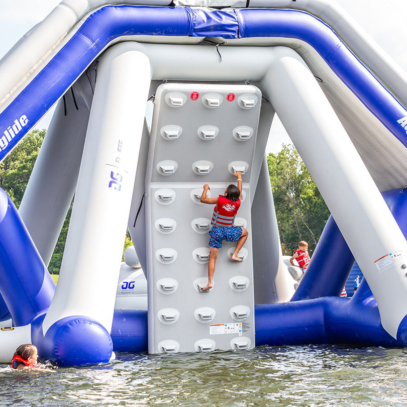 Child climbs El Jefe Climbing Wall at aquapark, others join the fun nearby.