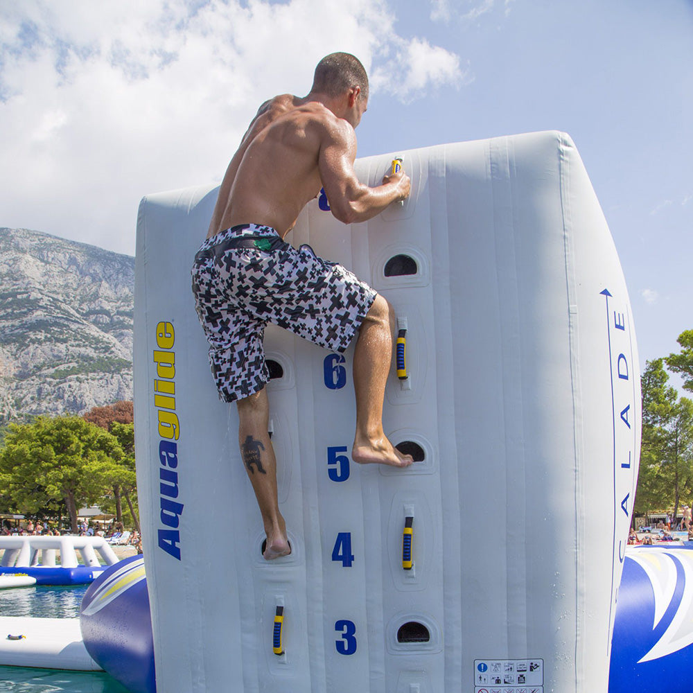 Man in Escalade 3M climbs inflatable wall on water with trees and mountains nearby.