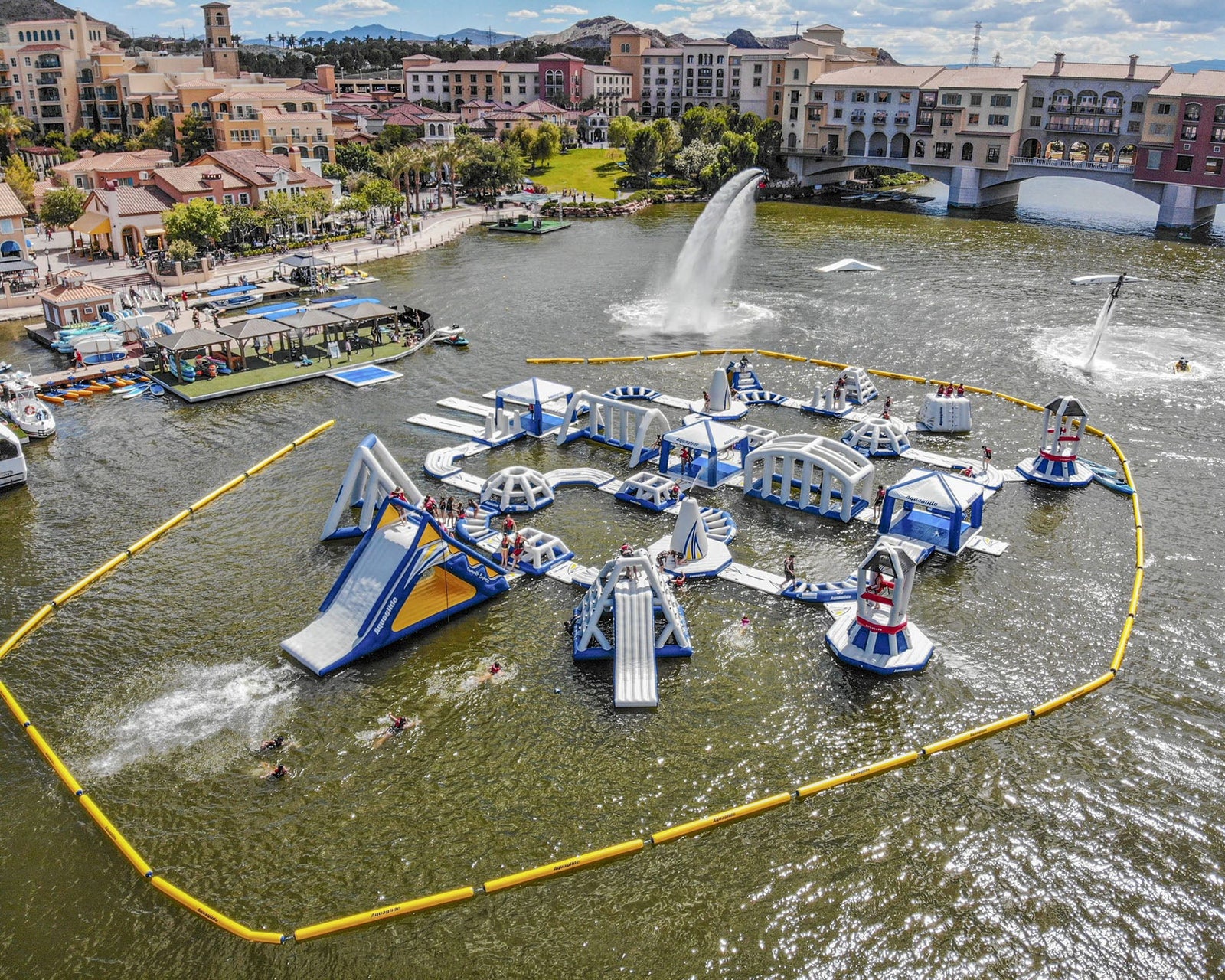 Floating inflatable water park with slides and obstacles on a lake, surrounded by buildings.