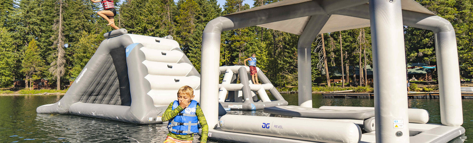Kids enjoying a floating inflatable water park on a lake surrounded by trees.