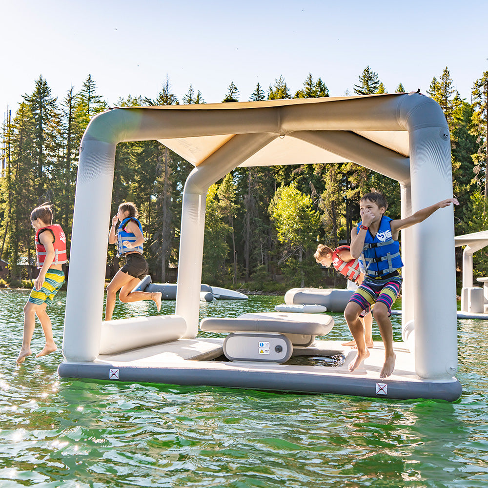 Kids in life vests leap from a Revel Lounge platform at an Aquaglide park amid trees.