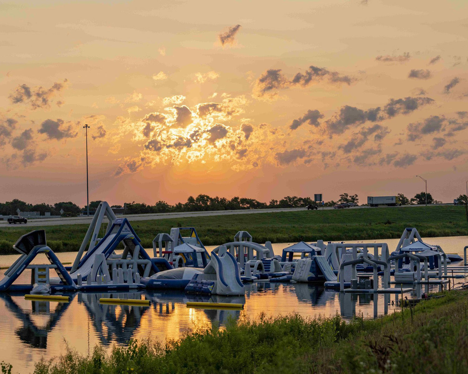 Sunset over an inflatable water park on a calm lake, with a highway and vehicles in the background.