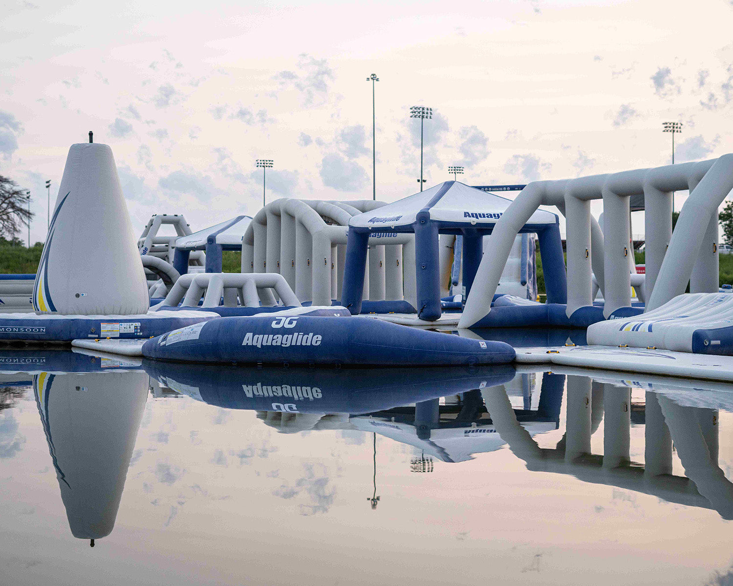 Inflatable water park structures mirrored in calm water under a cloudy sky.