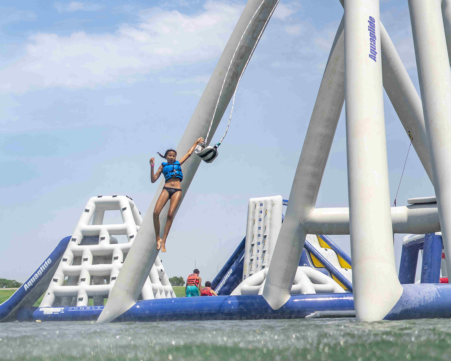 Person swinging on a rope at a water park with inflatable structures in the background.