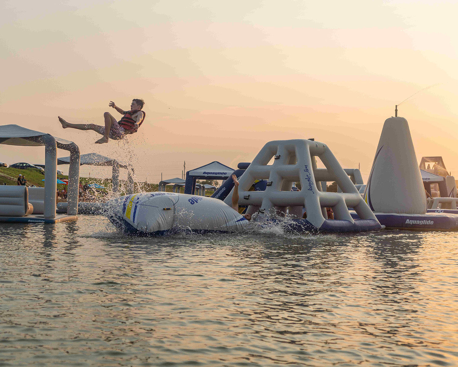 Person in life vest bouncing off inflatable water obstacle course at sunset.