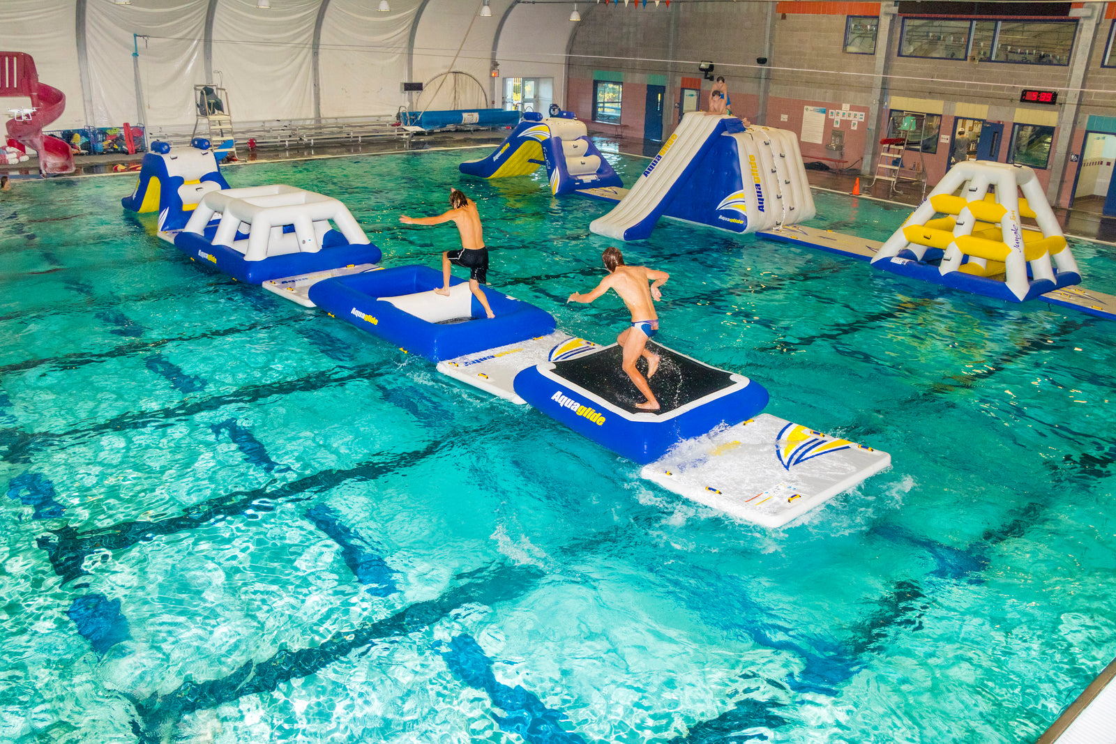 Two people balancing on an inflatable obstacle course in a large indoor pool.