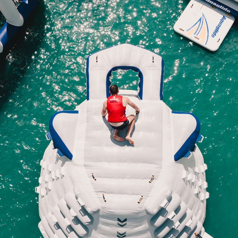Man wearing a red life jacket sitting on top of Aquaglide inflatable climbing wall and jump platform looking into the slide port.