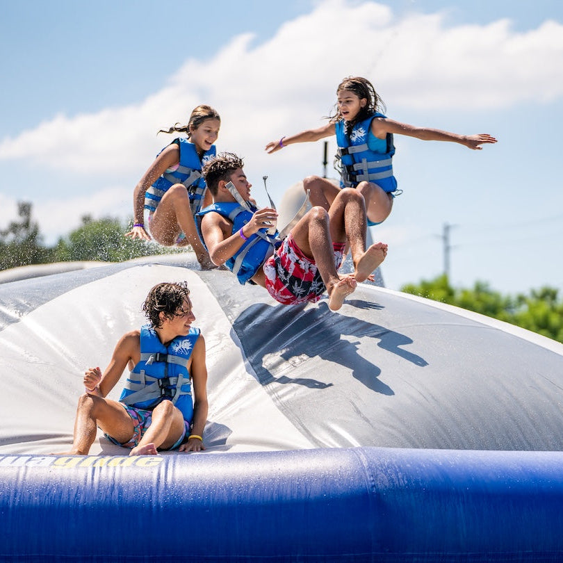 Kids in life vests bounce and slide on the Kaos inflatable at a water park on a sunny day.