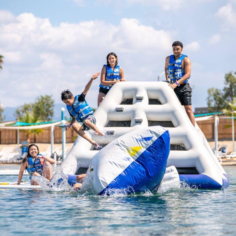 Group of kids and adults playing on Jungle Jim inflatable water structure in a lake.