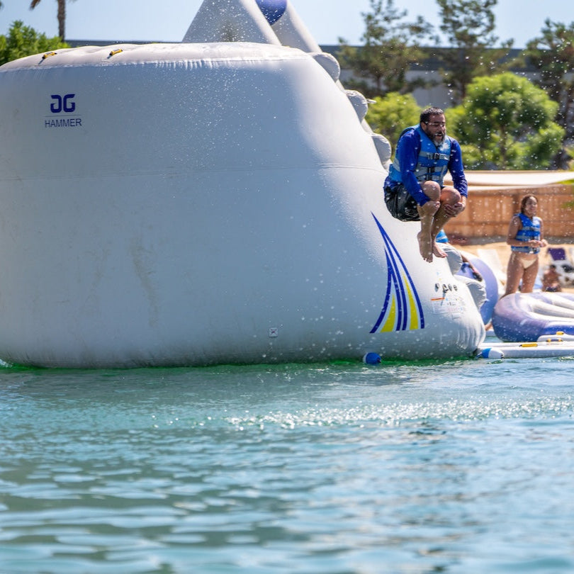 Adult jumping off the Aquaglide Hammer doing a cannonball into the water wearing a blue life jacket.