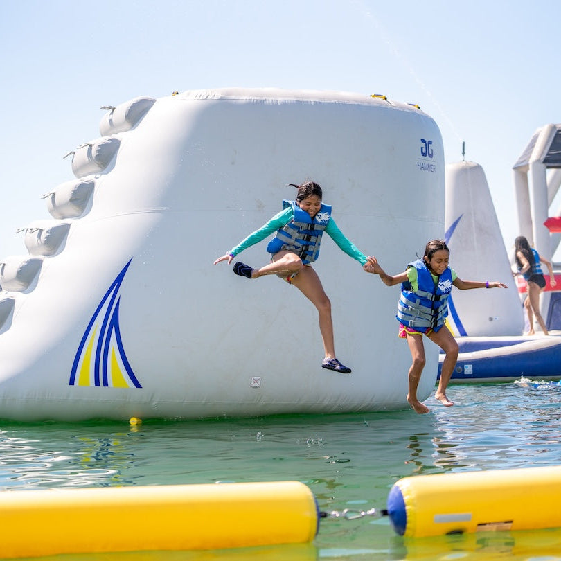 Two children in blue life jackets about to splash into the water after jumping off the Aquaglide Hammer.