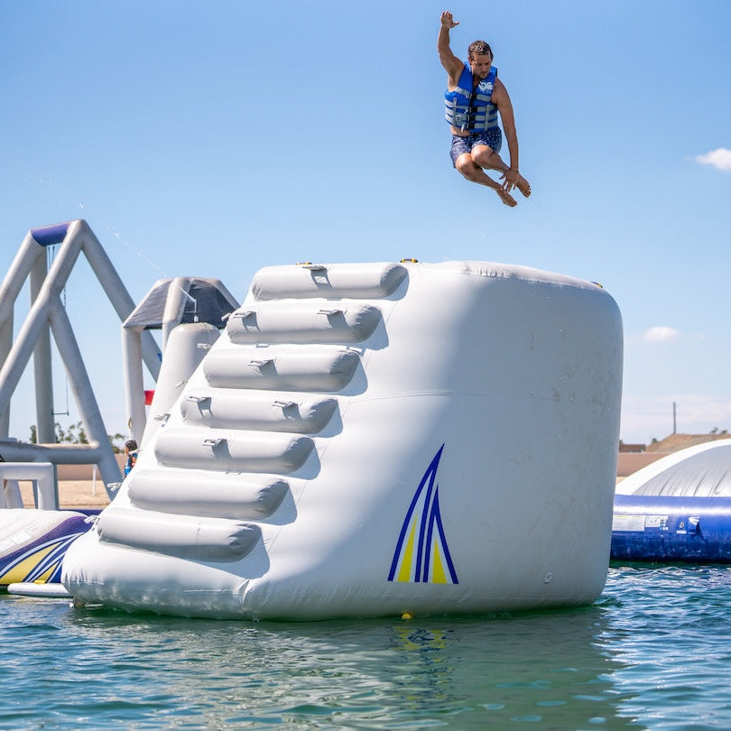 Man wearing a blue life jacket jumping off of the Aquaglide Hammer inflatable jump platform striking a pose mid air.