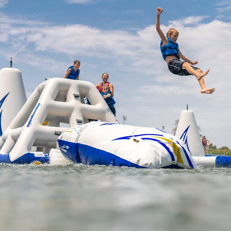 Child wearing a blue life jacket being launched into the air off of the Aquaglide Launch Bag with his family watching behind.
