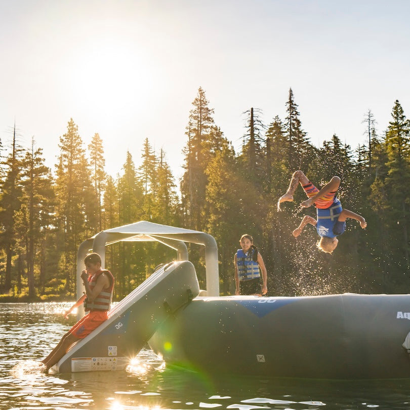 Young boy mid air doing a backflip on the Recoil 14 floating trampoline while another boy is sliding down the inflatable slide into the water.