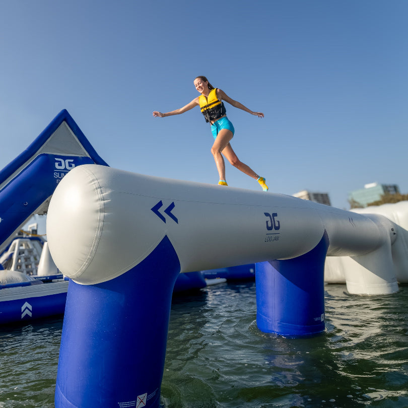 Person balancing on inflatable Log Jam obstacle over water with city skyline in background.
