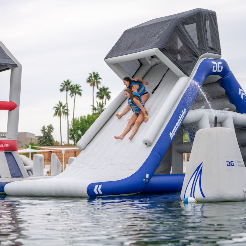 Two swimmers sliding down an Aquaglide Freefall Supreme lakefront slide. Ready to hit the water and holding hands.
