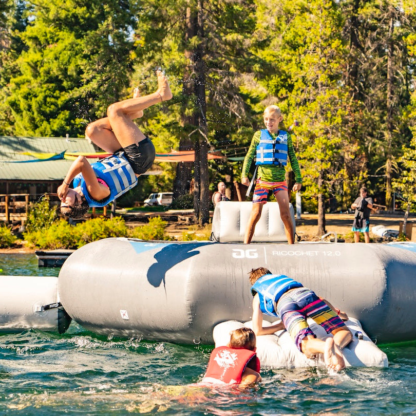 One child wearing a blue life jacket back flipping off of the Ricochet 12 floating trampoline while 3 other kids are playing and climbing out of the lake.