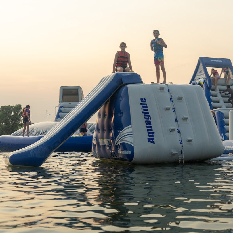 Two swimmers on top of the Aquaglide inflatable Aquapark and slide wearing a red and blue life jacket posing in the sunset.