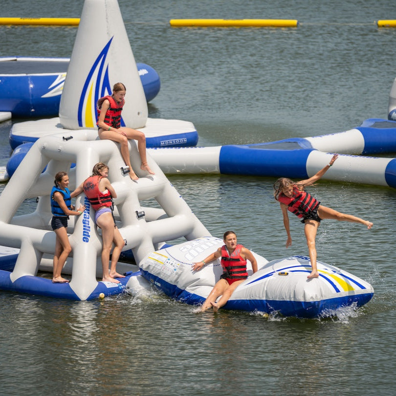 5 Swimmers wearing red and blue life jackets climbing on top of the Jungle Joe inflatable structure while one swimmer is in the air being launched off the Aquaglide Launch pad into the water.