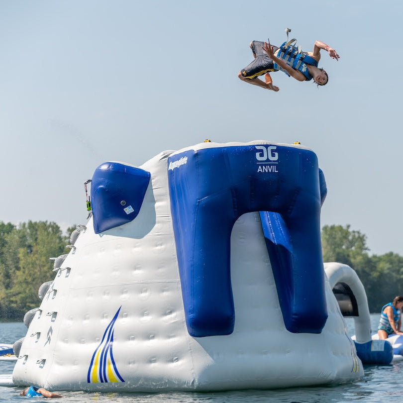 Kid wearing a blue life jacket backflipping off the top platform into the lake.