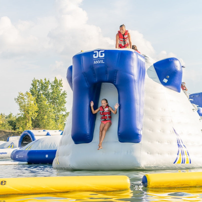 Young girl sliding down the slide port of the Aquaglide Anvil while another swimmer wearing a red life jacket watches from above.