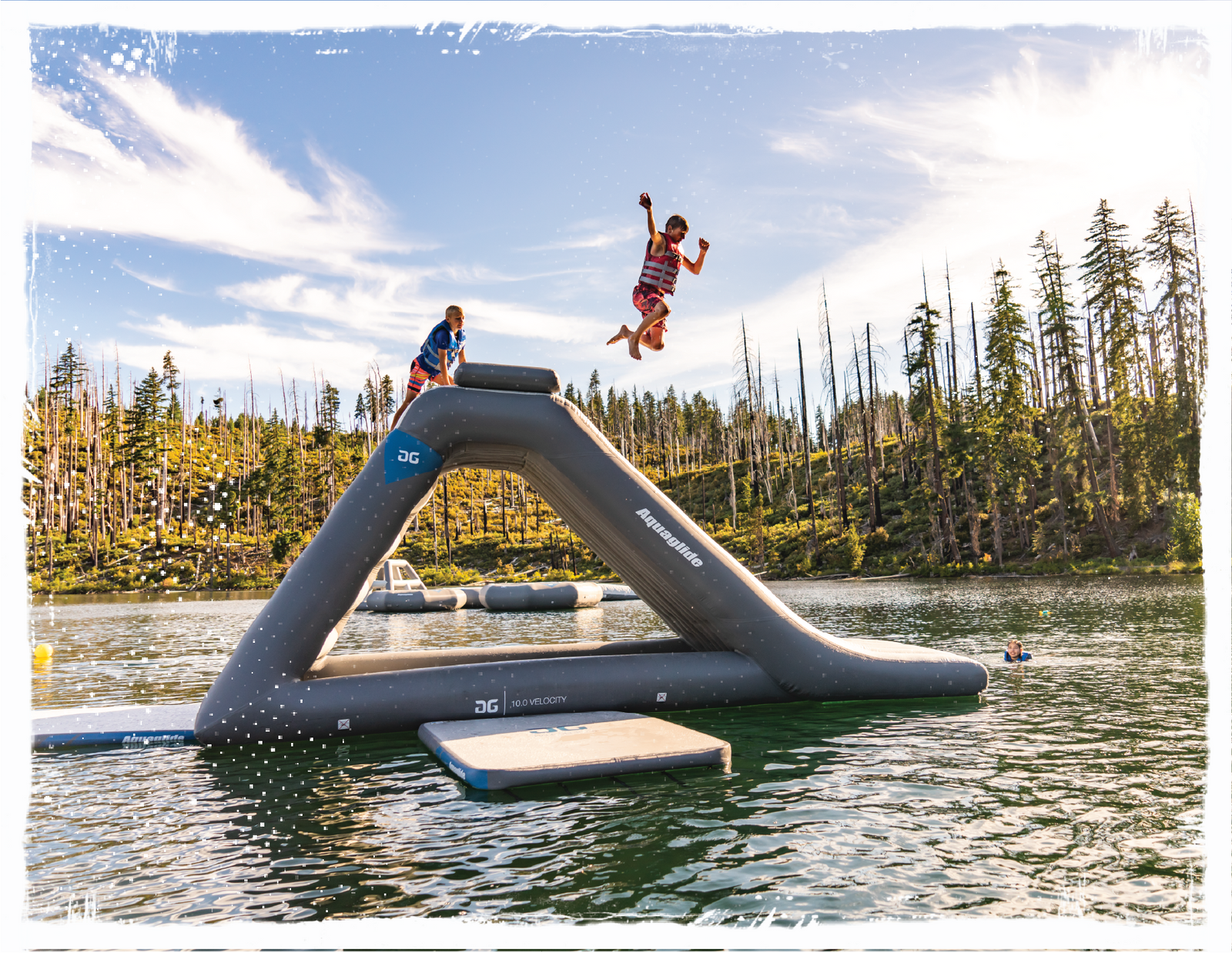 Child jumping off the top of gray Aquaglide floating slide and deck with alpine trees in the background. 